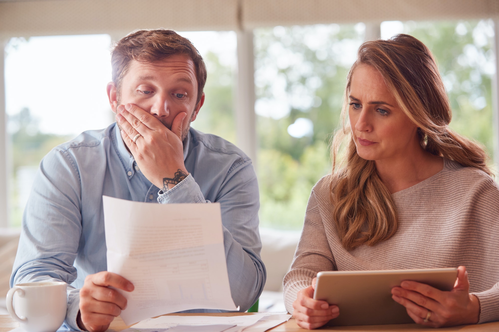 Worried Couple With Bills And Digital Tablet Sitting At Table At Home Reviewing Domestic Finances